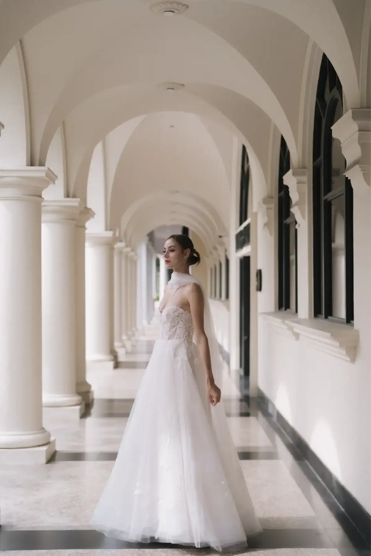 A woman in a white wedding dress stands in a grand, arched corridor with tall columns, soft natural light, and a calm, elegant atmosphere.