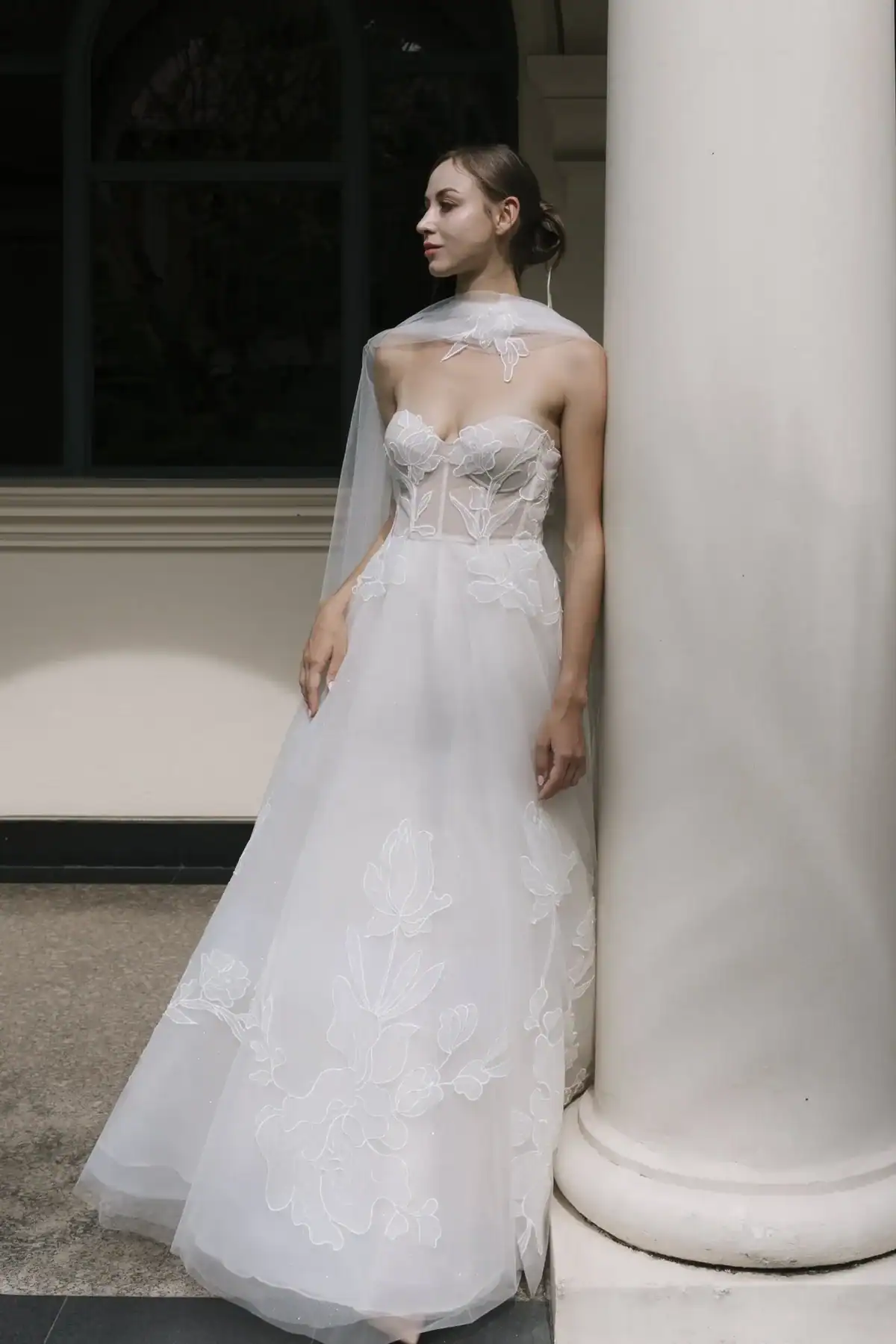 A woman in a strapless, white, floral-embroidered wedding dress stands against a white column. Her dress features sheer fabric and a flowing skirt. She gazes to the side, with a sheer veil draped around her neck and shoulders.