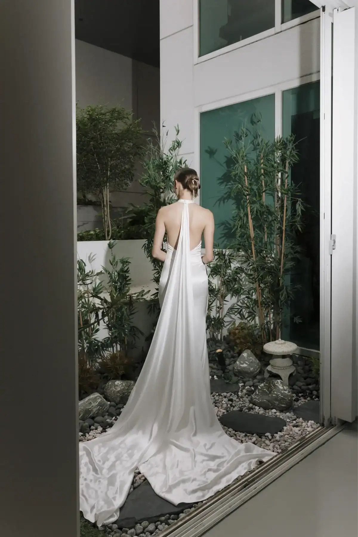 A woman in a long, backless white gown with a flowing train stands in a modern, minimalist garden with green plants and rocks, facing away from the camera.