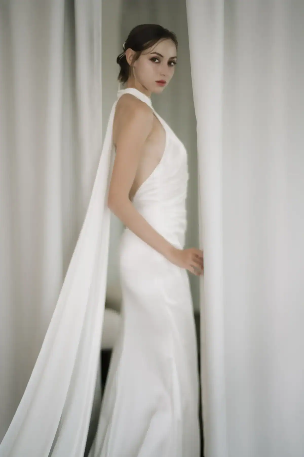 A woman in a sleek, white, halter-neck gown stands between white curtains, looking over her shoulder with a serious expression. The dress features a long, flowing cape. The setting is softly lit and elegant.