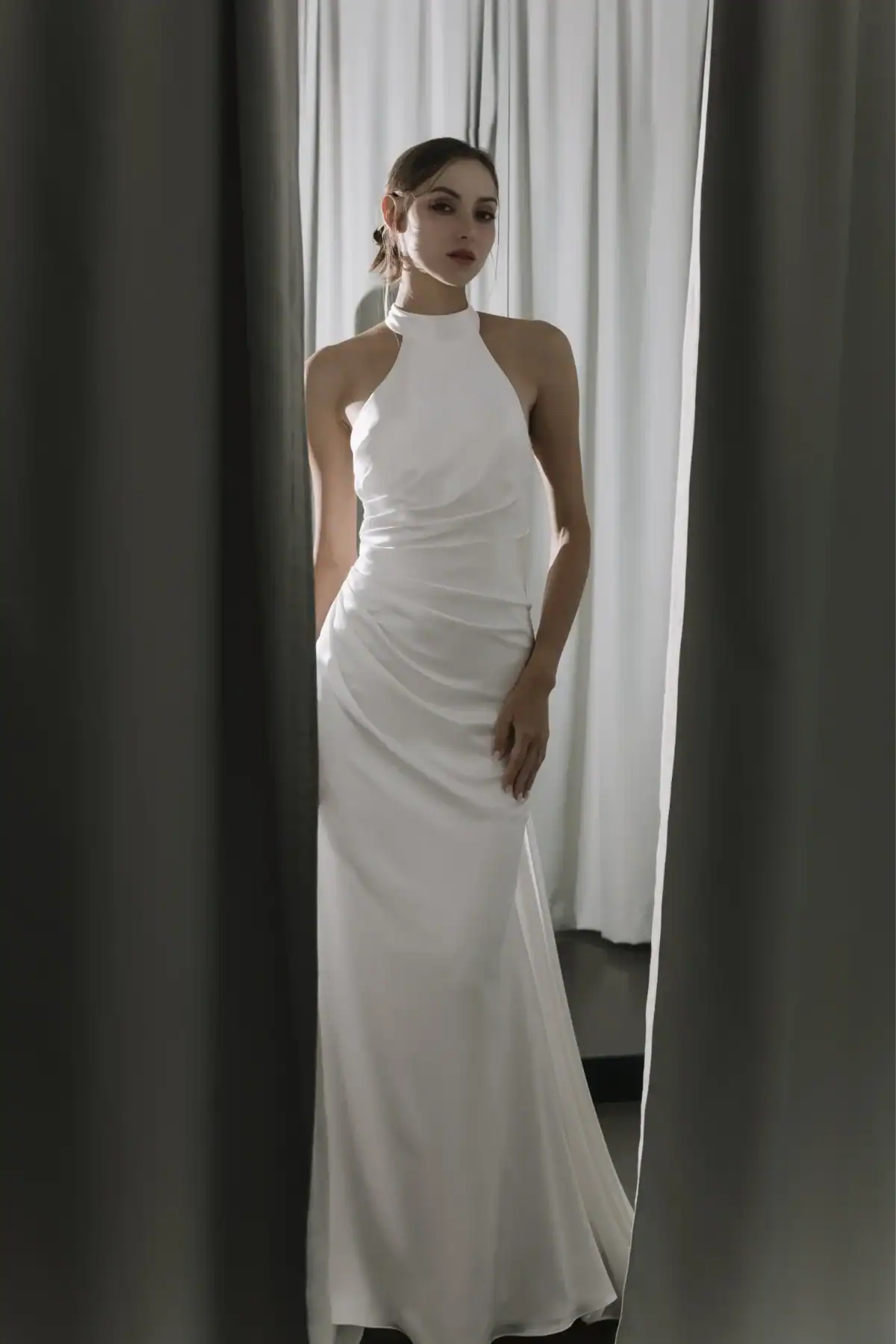 A woman in a sleeveless, high-neck white gown stands between parted grey curtains, looking at the camera with a neutral expression. The background features white drapes and soft lighting.