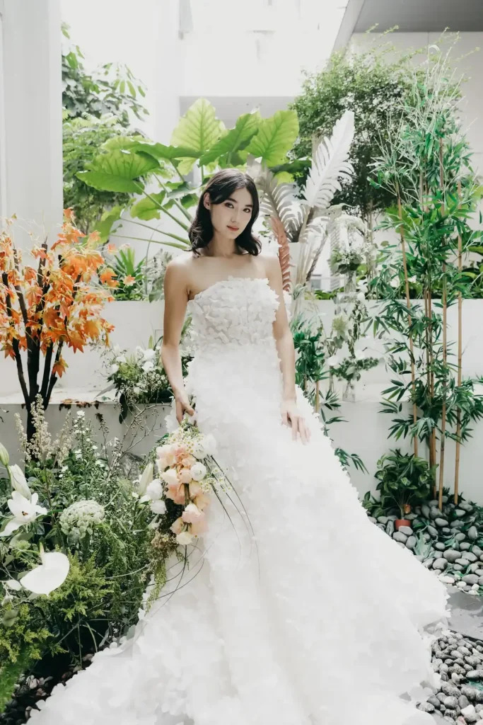 A woman in a strapless white wedding dress stands in a lush garden surrounded by green plants and orange flowers, holding a bouquet of peach and white roses, looking at the camera.