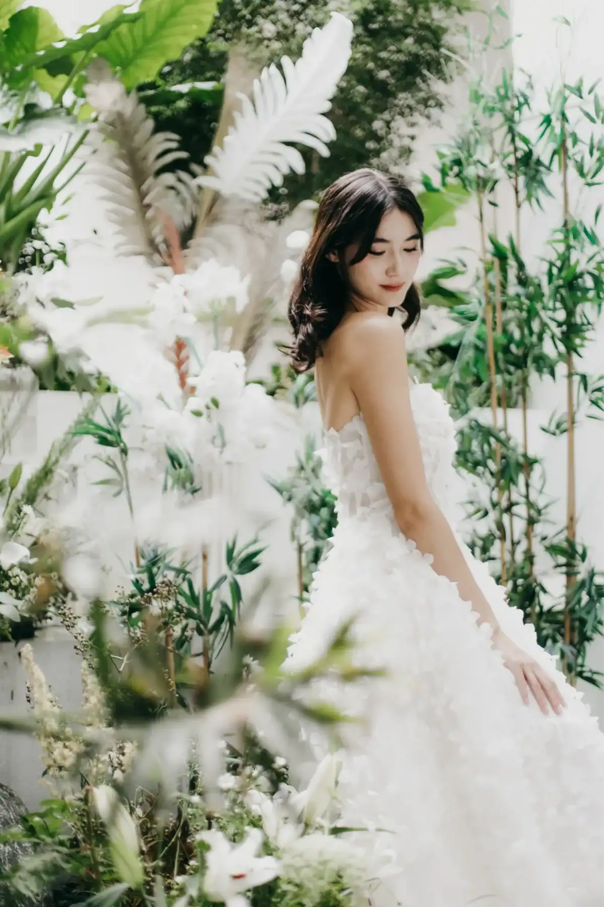 A woman in a strapless white dress stands gracefully among lush green plants and white flowers, looking down with a gentle expression. The setting is bright and filled with natural light.