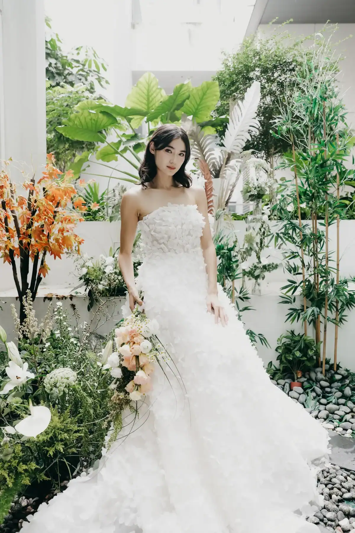 A woman in a strapless white wedding dress stands in a lush garden surrounded by green plants and orange flowers, holding a bouquet of peach and white roses, looking at the camera.