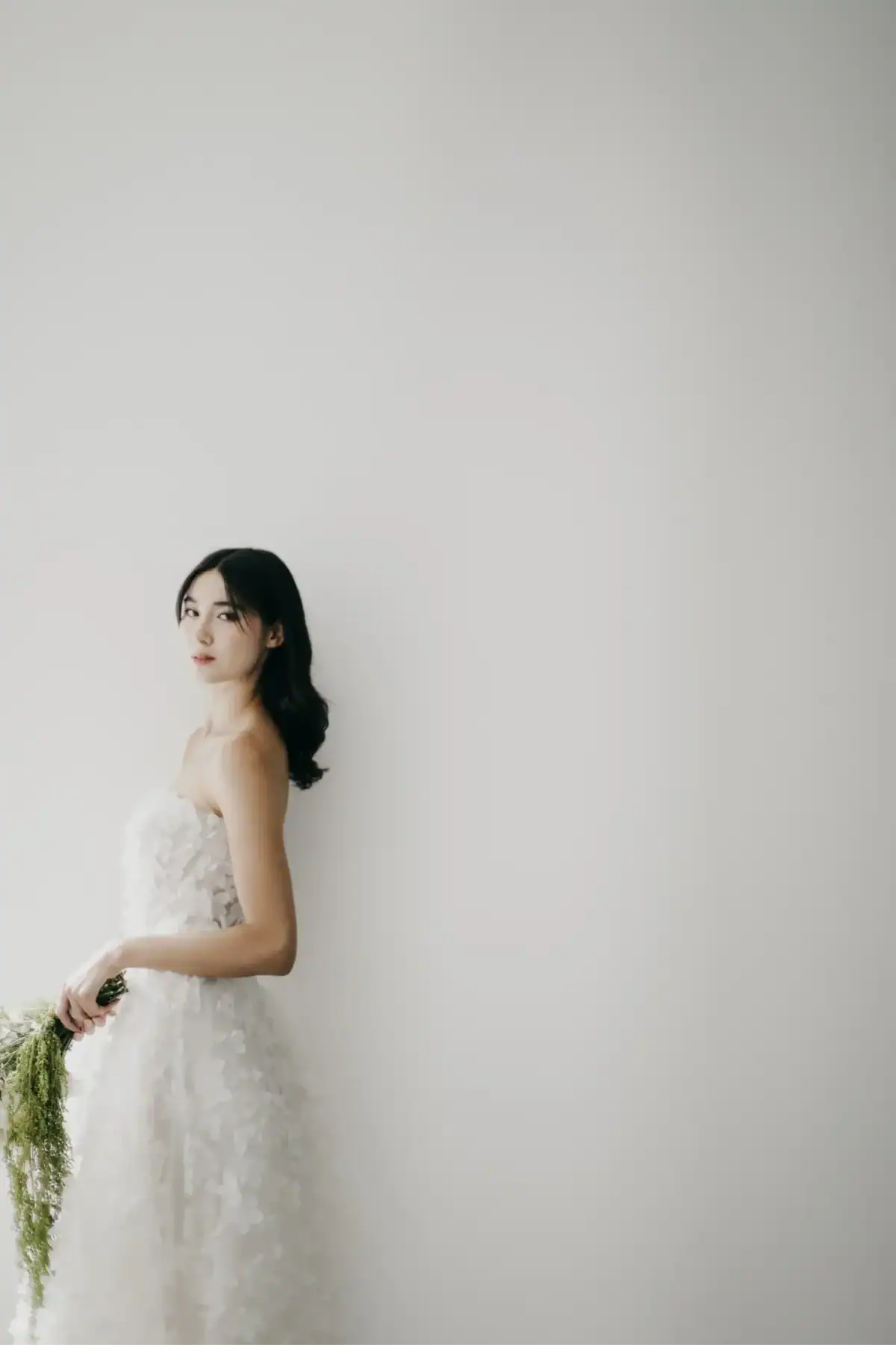 A woman in a strapless white dress holding green foliage stands against a plain, light-coloured background, gazing softly towards the camera.