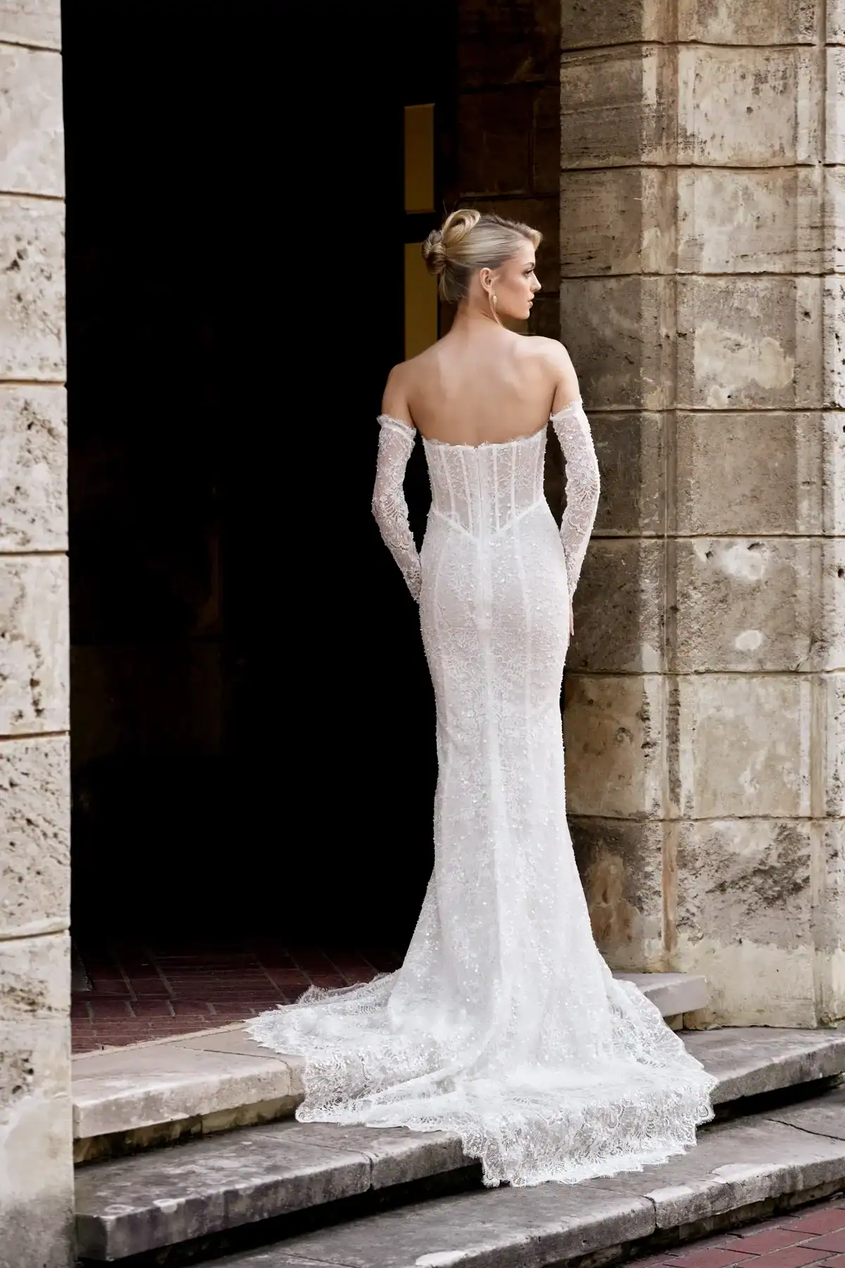 A woman in an elegant, off-the-shoulder, figure-hugging white wedding dress with a long train stands on stone steps in front of a rustic stone building, facing away from the camera.