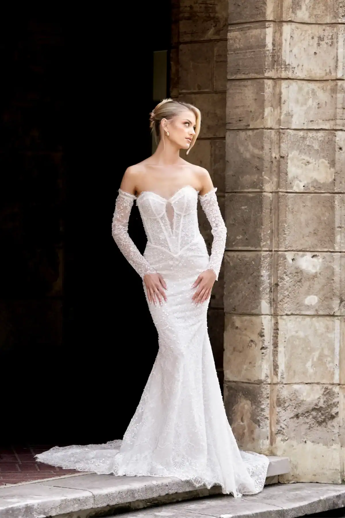 A woman in an off-the-shoulder, figure-hugging white lace wedding dress stands by a stone wall, looking to the side with her hands resting on her hips.
