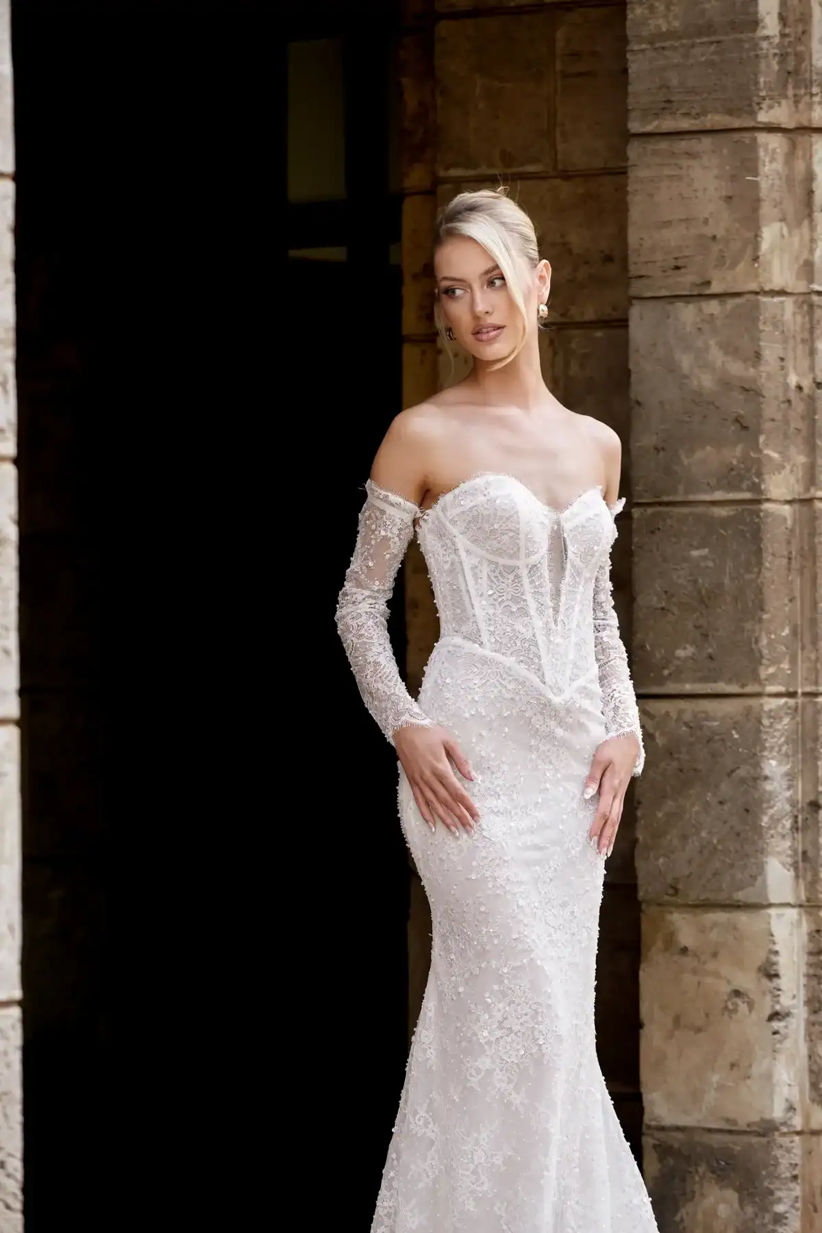 A woman wearing a fitted, off-the-shoulder, white lace wedding dress with sheer long sleeves stands beside a stone wall, looking slightly to the side.