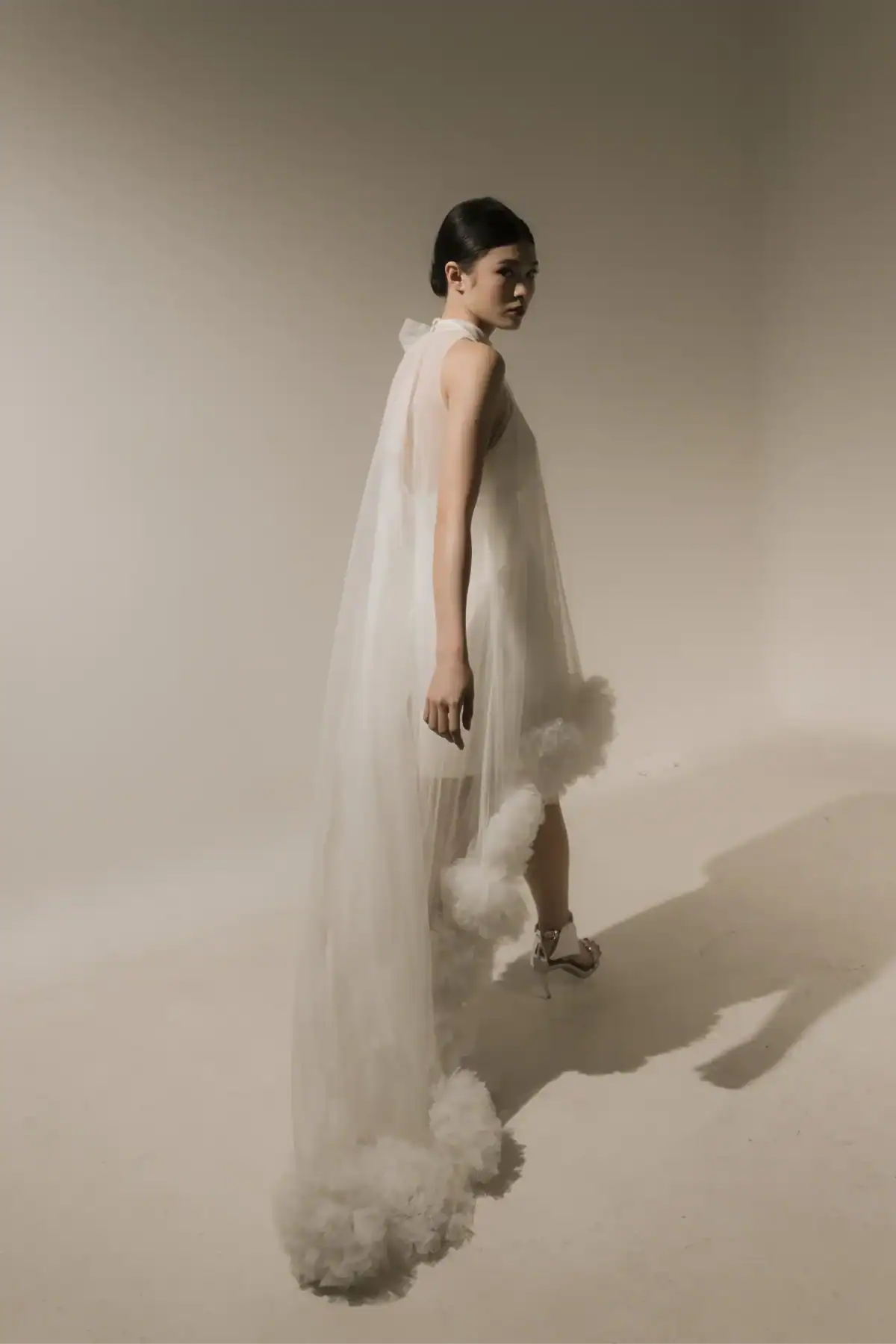 A woman stands in a minimalist studio wearing a sheer white gown with a long tulle train adorned with fluffy white accents, paired with strappy heels. She looks over her shoulder, softly lit by natural light.