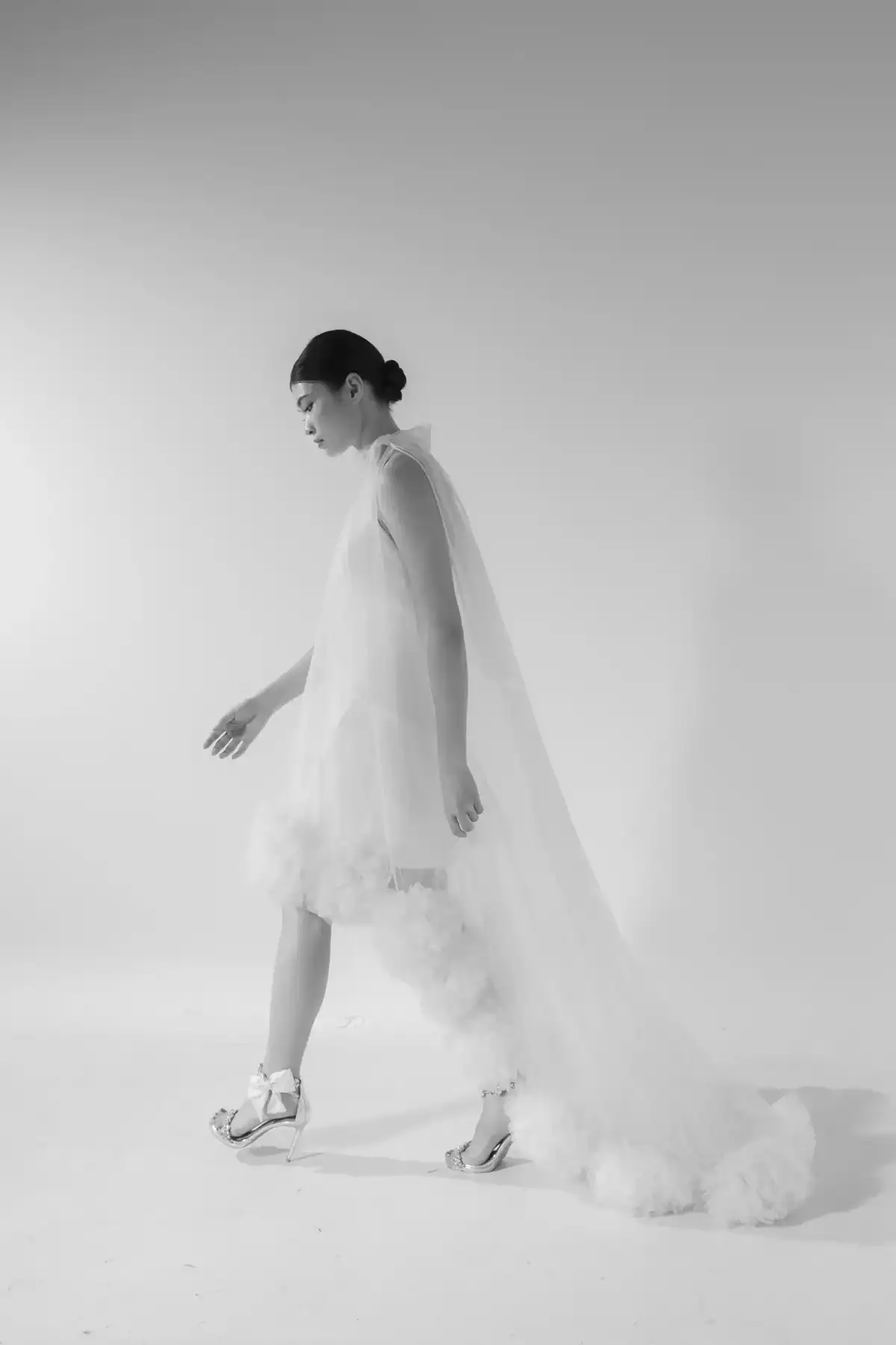 A woman in a long, sheer, white dress with a voluminous, tulle train walks sideways in a minimalist studio. She has her hair in a low bun and is wearing white, strappy platform shoes. The image is in black and white.