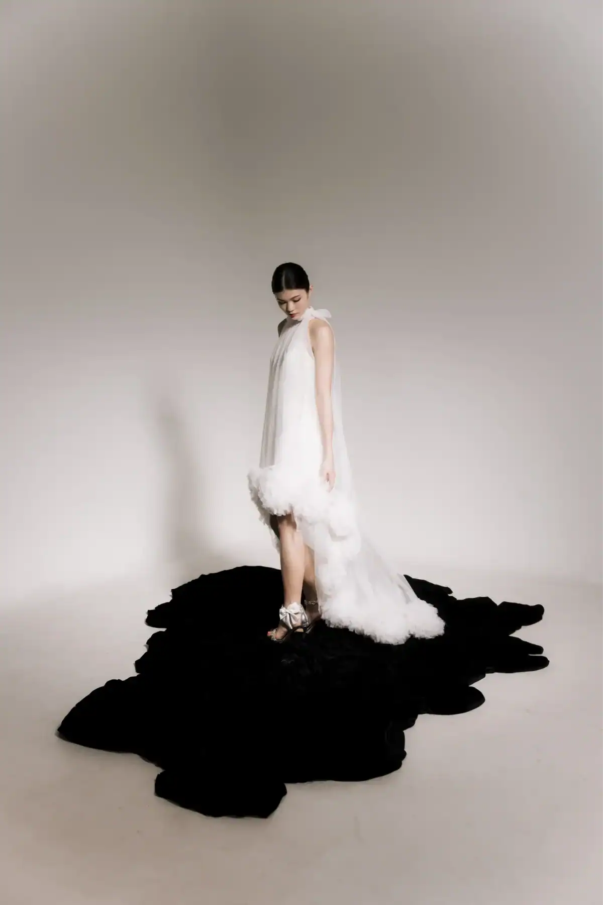 A woman in a white, frilled high-low dress stands on an irregularly shaped black rug in a minimal, light-coloured room, looking down with her hair tied back and wearing silver shoes.