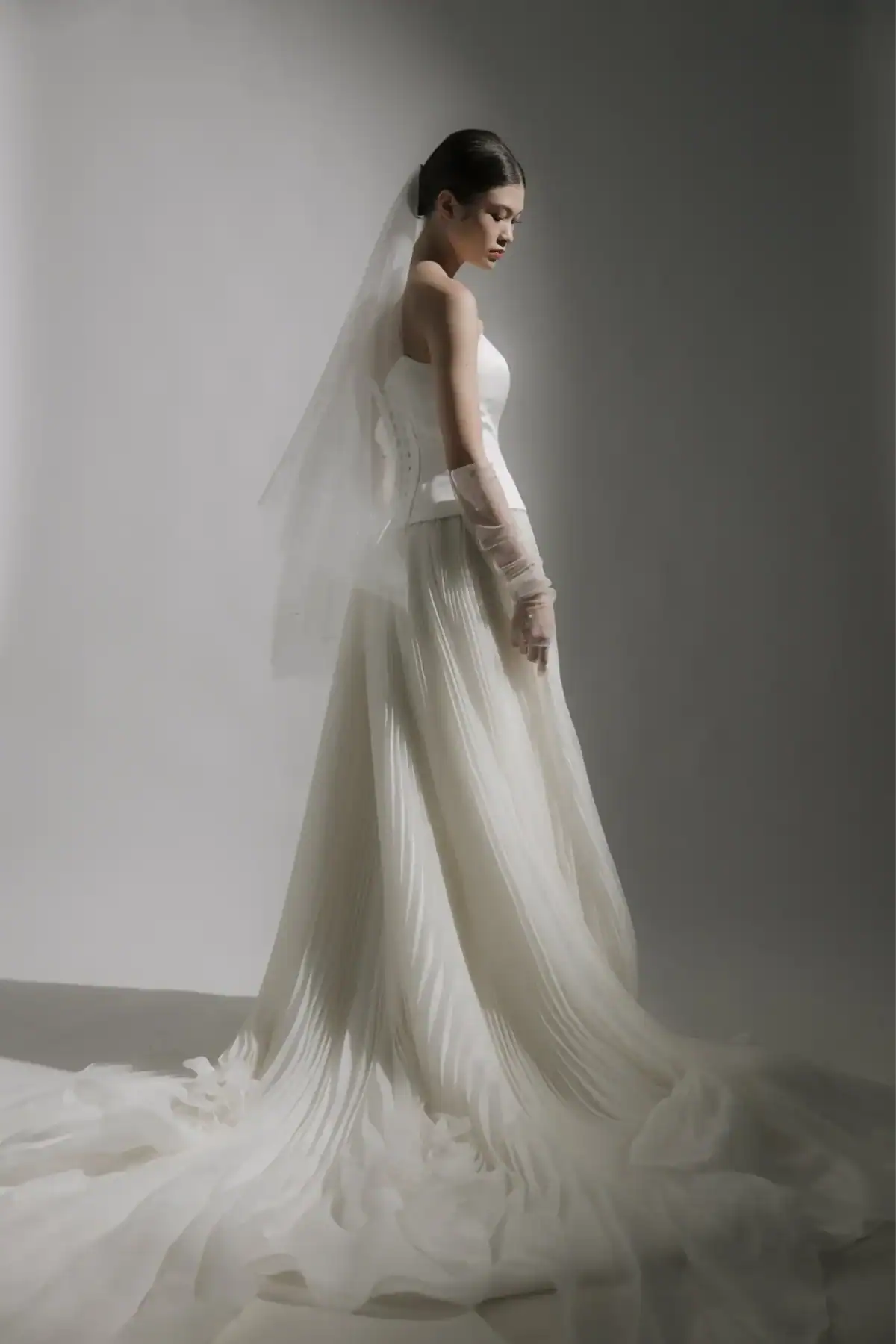A woman in a strapless white wedding dress with a long, pleated skirt and veil stands in profile against a neutral background, looking down softly with her hands by her side.