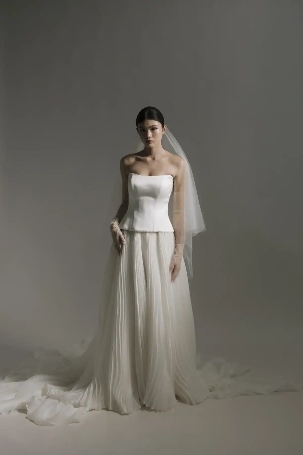 A woman in a strapless white wedding dress with a fitted bodice, flowing pleated skirt, and sheer veil stands against a plain grey background, looking slightly downward with her hands by her sides.