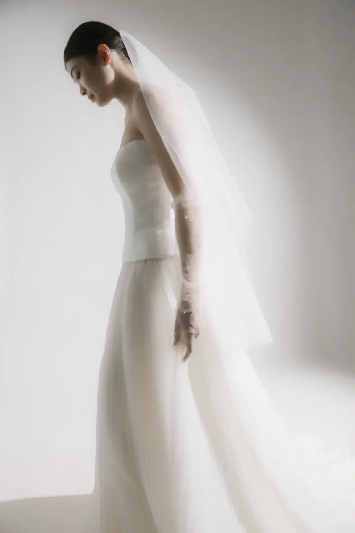 A woman in a strapless white wedding dress and long veil stands in profile against a soft, light background, looking downwards with her hands by her side.