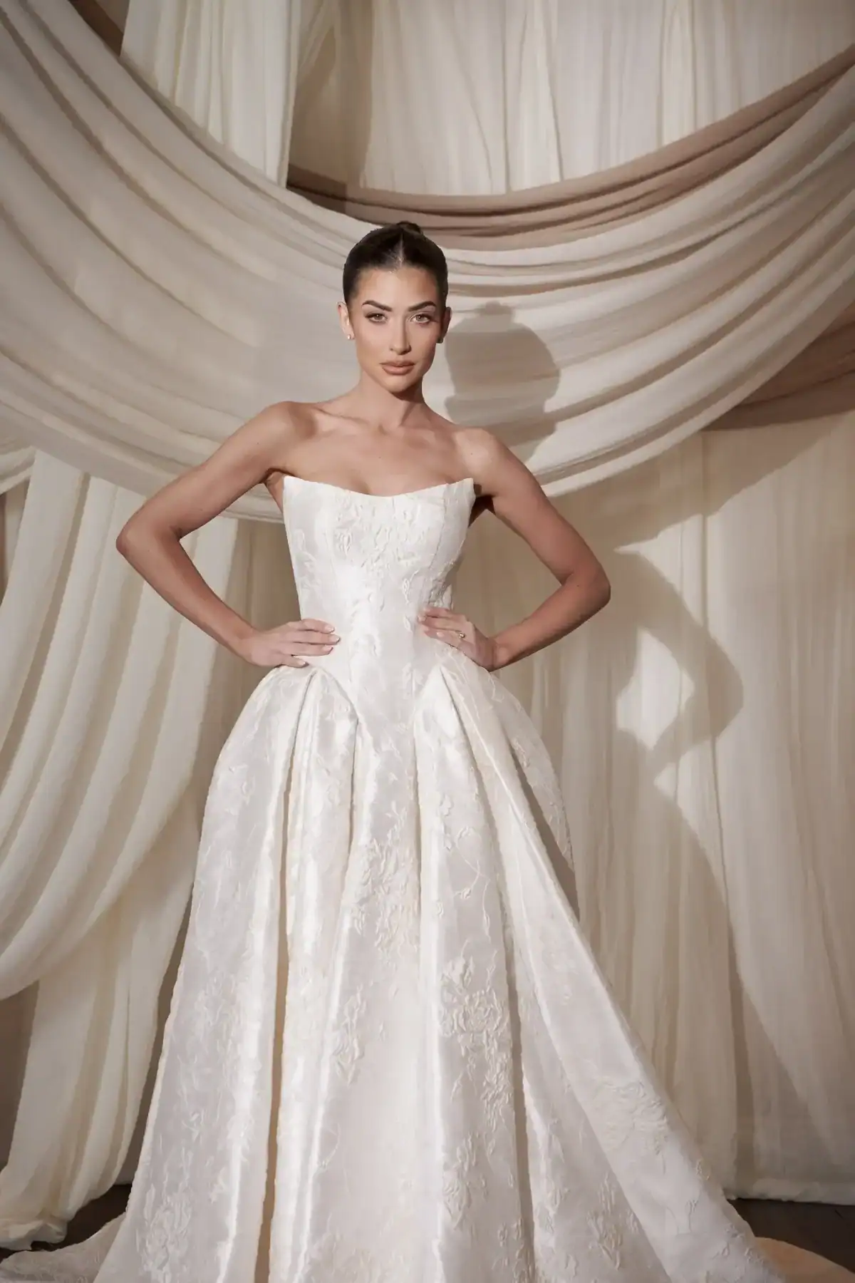 A woman in a strapless white bridal dress with a full skirt stands confidently with hands on hips in front of flowing cream-coloured curtains.