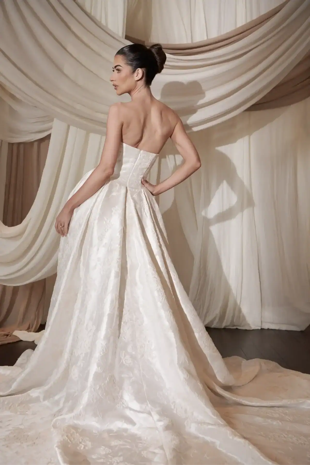 A woman stands in front of draped ivory fabric, wearing a strapless, floor-length wedding dress with a fitted bodice and a long, flowing train. She poses with one hand on her hip, showing the back of the dress.