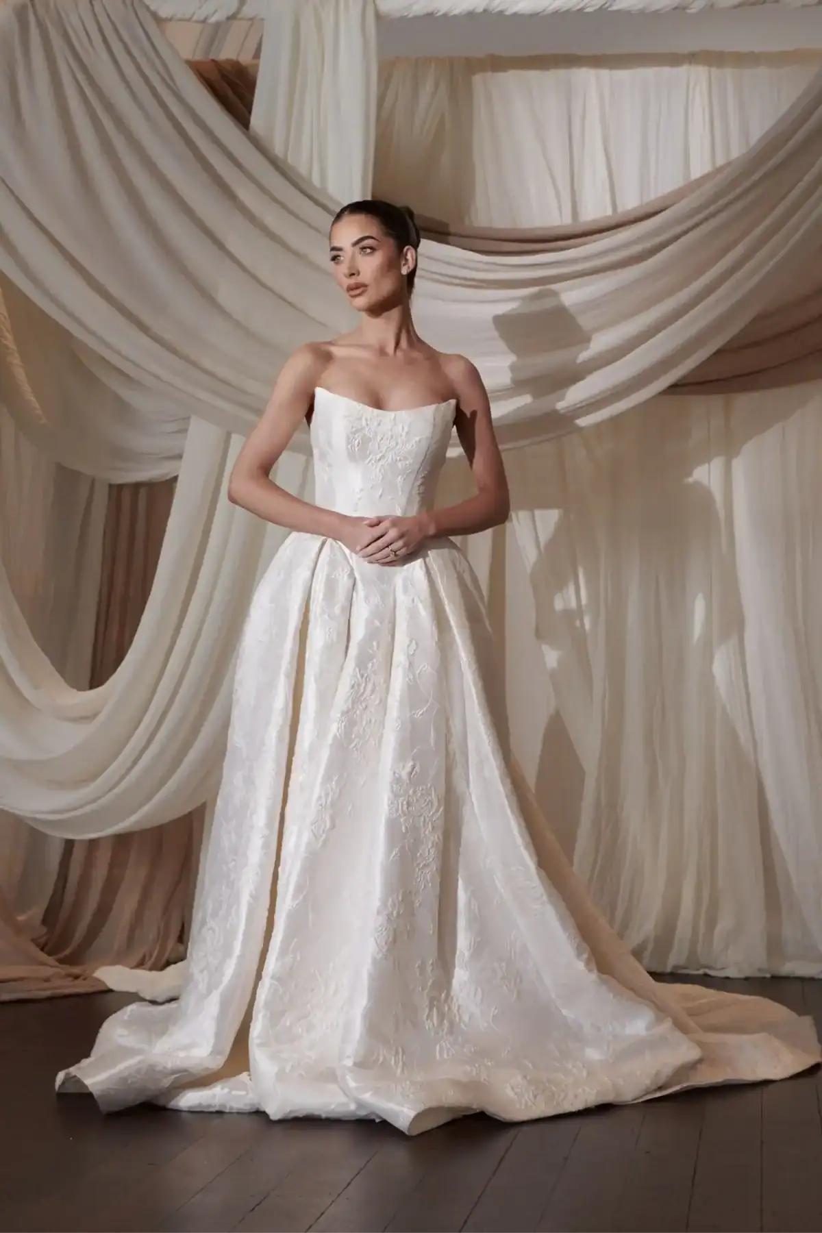 A woman in a strapless, white, embroidered ball gown stands with her hands clasped, posing in front of softly draped, cream-coloured fabric backdrops.