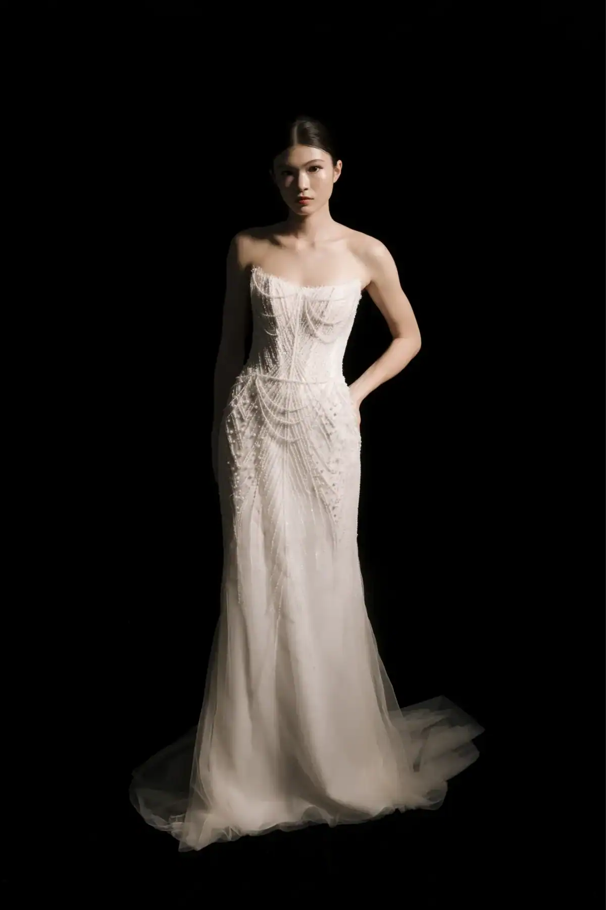 A woman stands against a black background wearing a strapless, fitted, white wedding dress with intricate lace detailing and sheer tulle at the bottom, with her hands in her pockets and a calm expression.