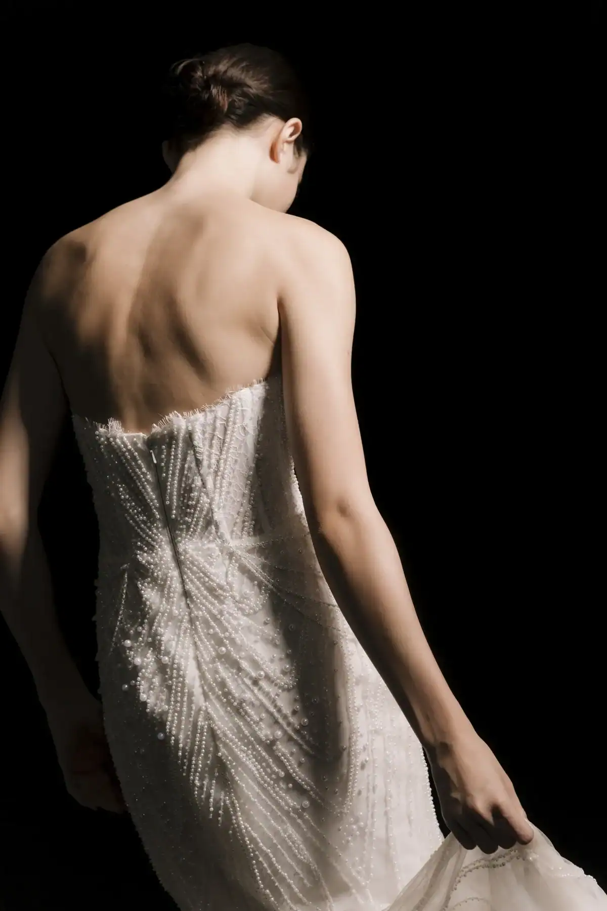 A woman in a strapless, beaded white dress stands with her back to the camera against a black background, holding part of her skirt and looking down. Her hair is styled in a low bun.