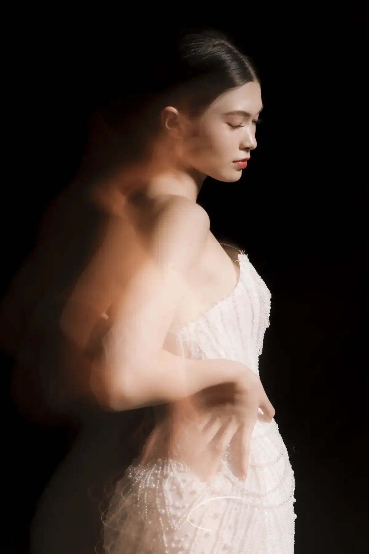 A woman in a white, beaded dress poses against a black background. She stands in profile with eyes closed, and her blurred arm creates an ethereal, double-exposure effect.
