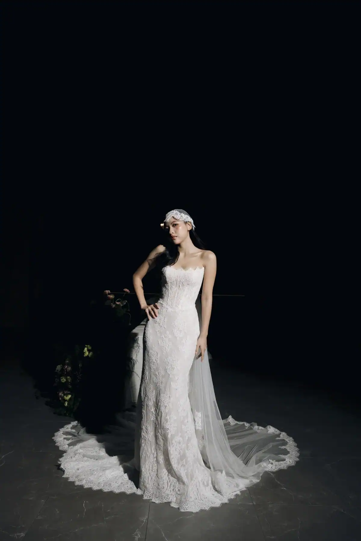 A woman in a white lace wedding dress with a long train and veil stands confidently against a dark background, one hand on her hip.
