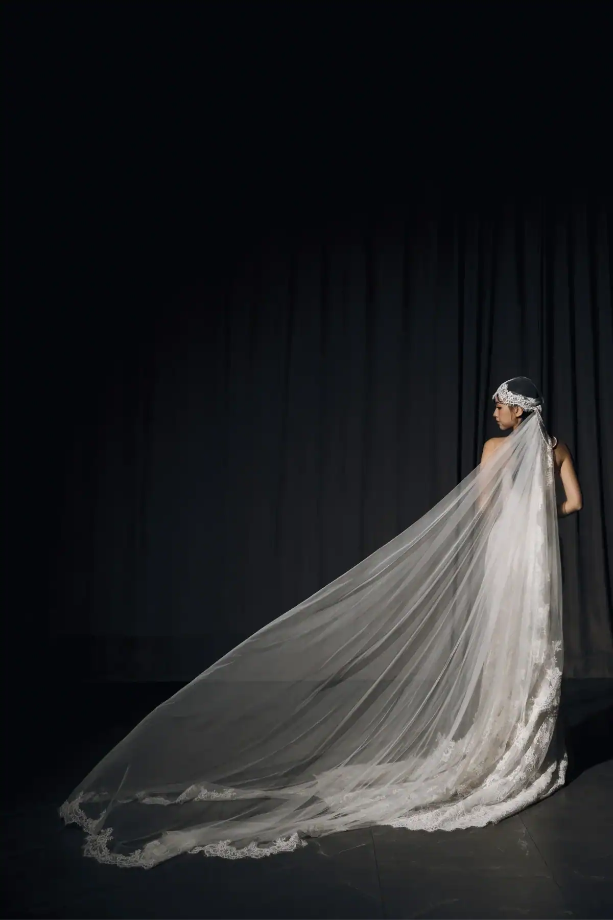 A bride in a white dress with a long, flowing lace veil stands facing away against a dark backdrop, looking to the side, illuminated softly from the right.