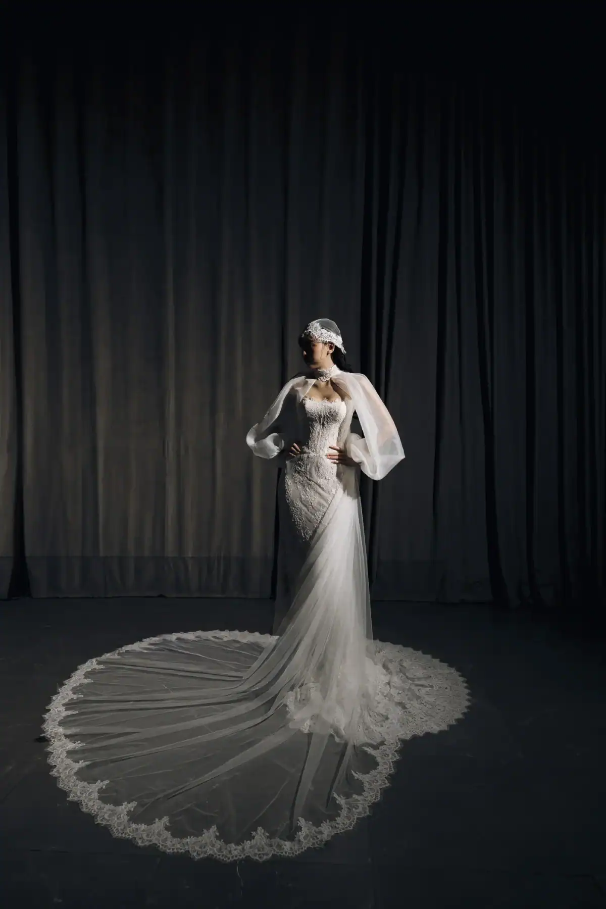 A woman in an elegant, fitted wedding dress with a long lace train and sheer sleeves stands confidently against a dark curtain backdrop, wearing a decorative headpiece.