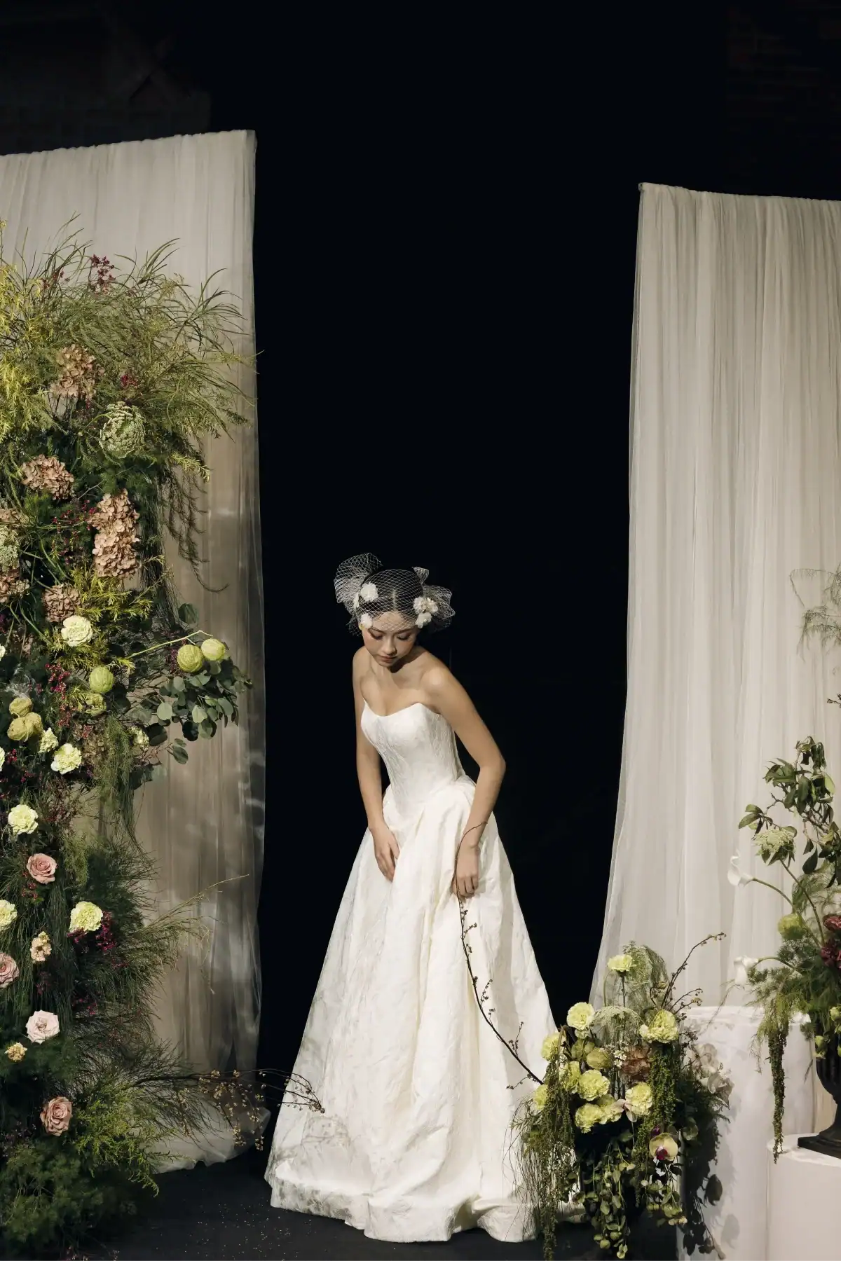 A woman in a strapless white wedding dress and birdcage veil stands between sheer curtains, surrounded by lush floral arrangements and greenery.