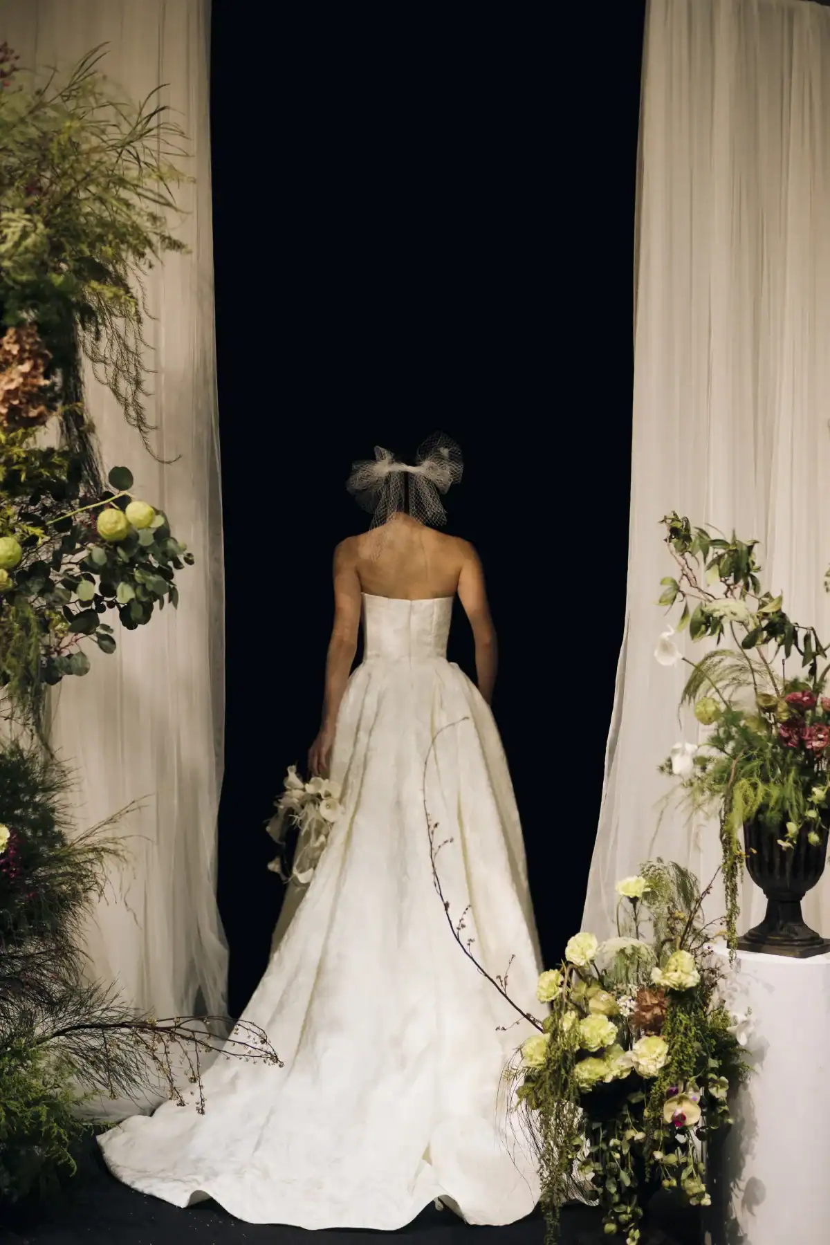 A bride in a strapless white gown and veil stands facing away between white curtains, surrounded by lush green and white floral arrangements.