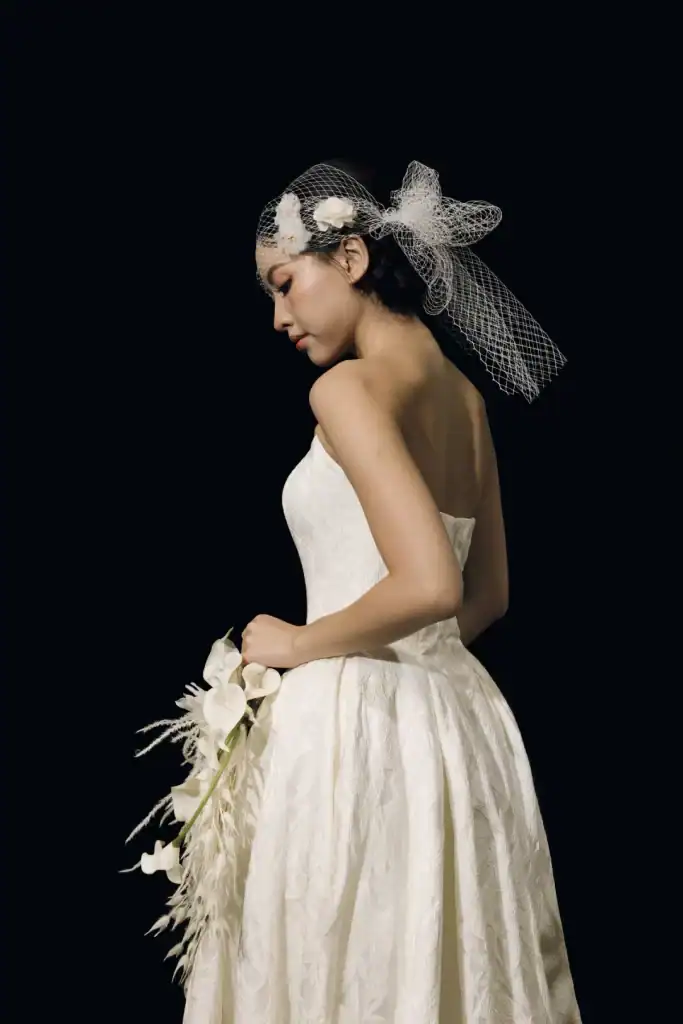 A woman in a white strapless wedding dress and veil holds a bouquet of white flowers, standing in profile against a black background.