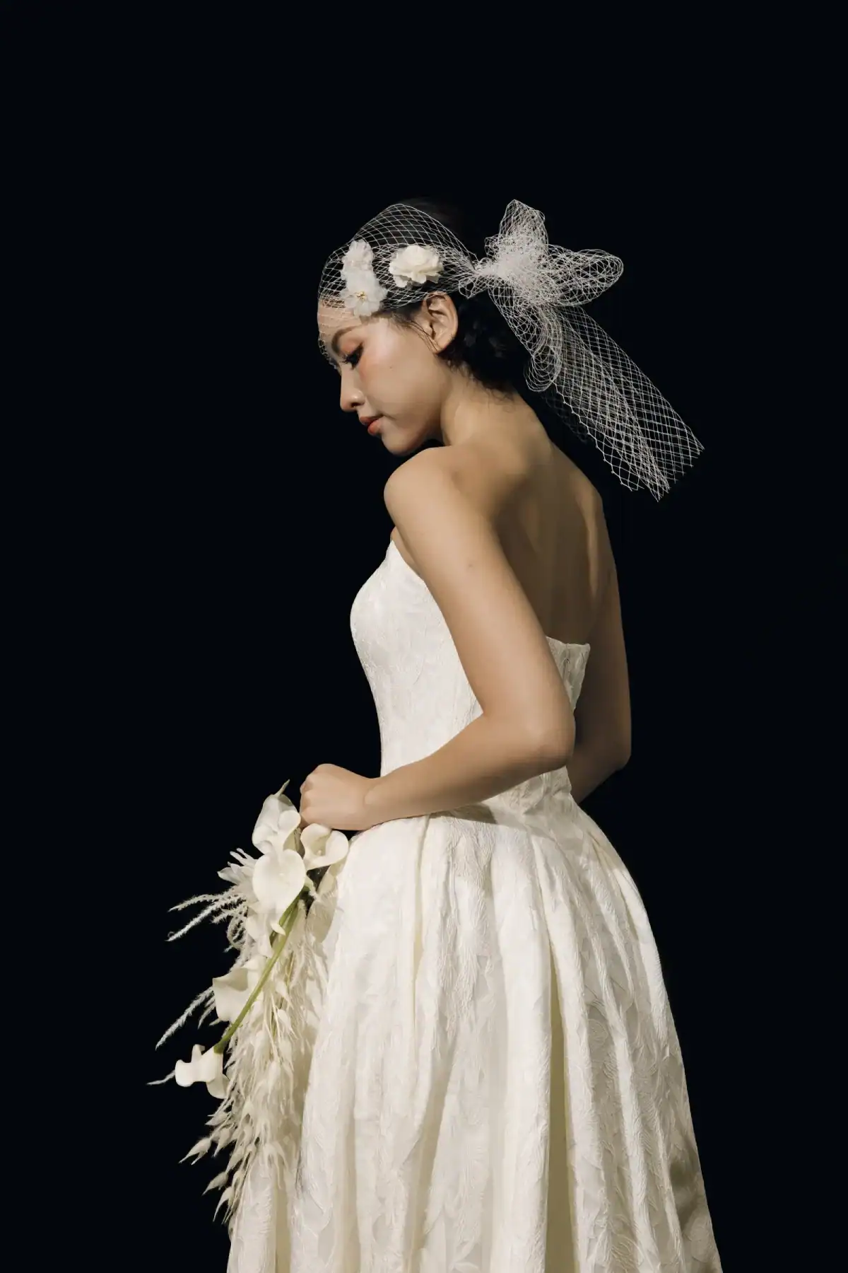 A woman in a white strapless wedding dress and veil holds a bouquet of white flowers, standing in profile against a black background.