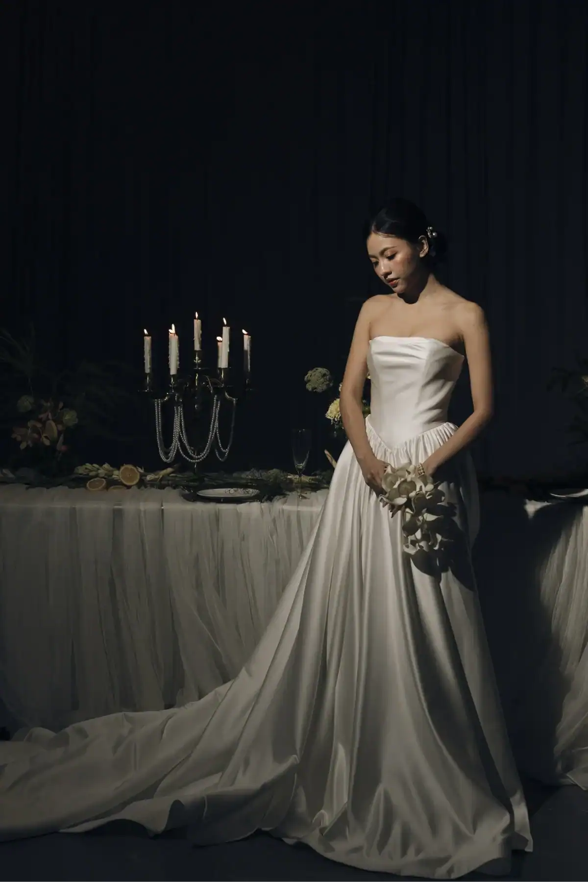 A bride in a white strapless gown stands by a candlelit table with a white tablecloth, holding a bouquet, looking down in a dimly lit, elegant setting.