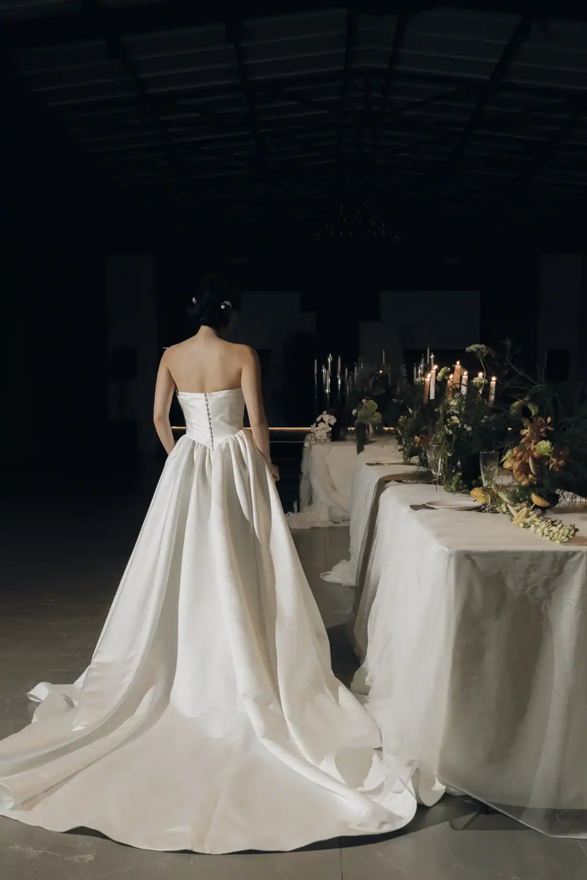 A bride in a white dress stands facing a candlelit wedding reception table adorned with flowers, seen from behind in a dimly lit room.