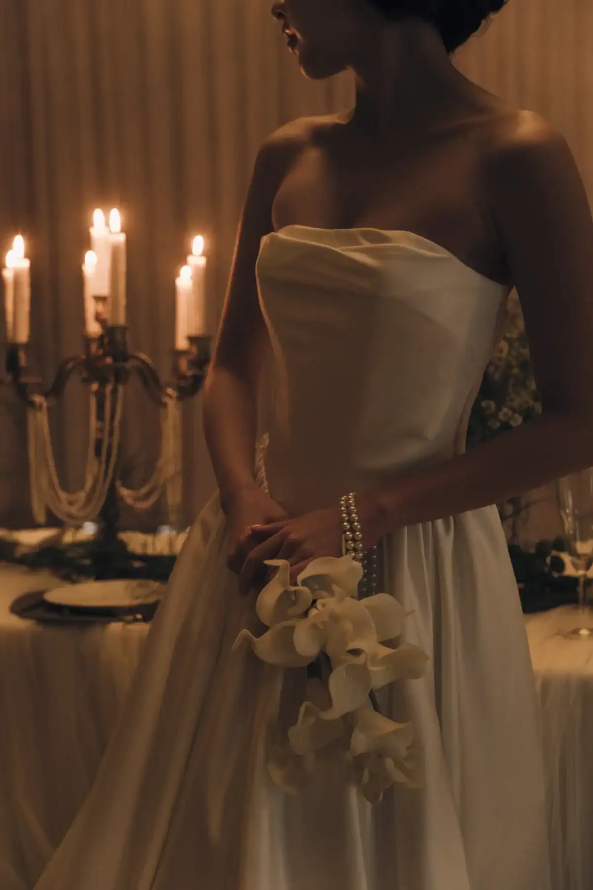 A woman in a strapless white wedding dress holds white calla lilies and a pearl bracelet, standing near a candlelit table with a candelabrum in the background.