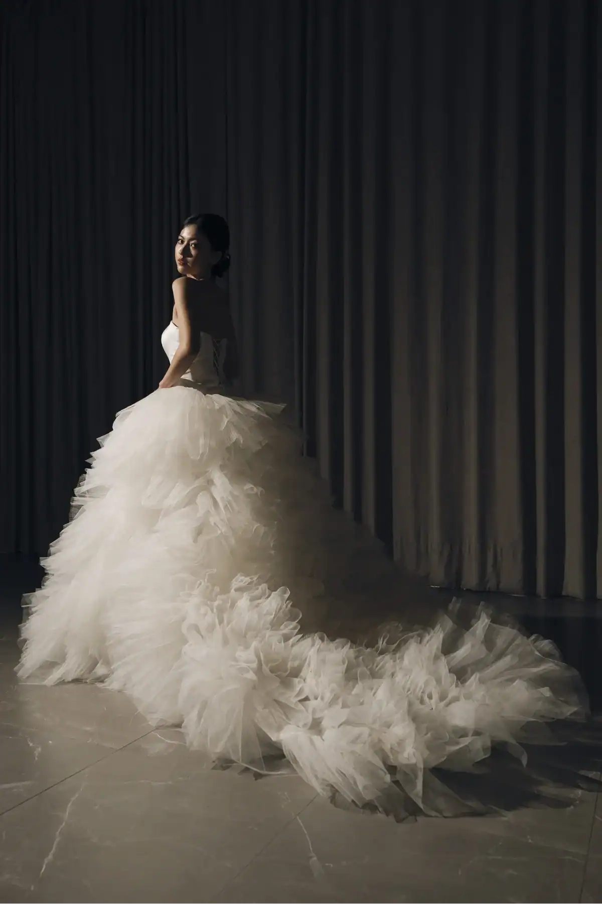 A woman in a strapless, voluminous white wedding dress stands on a marble floor, looking over her shoulder with soft lighting and dark curtains in the background.