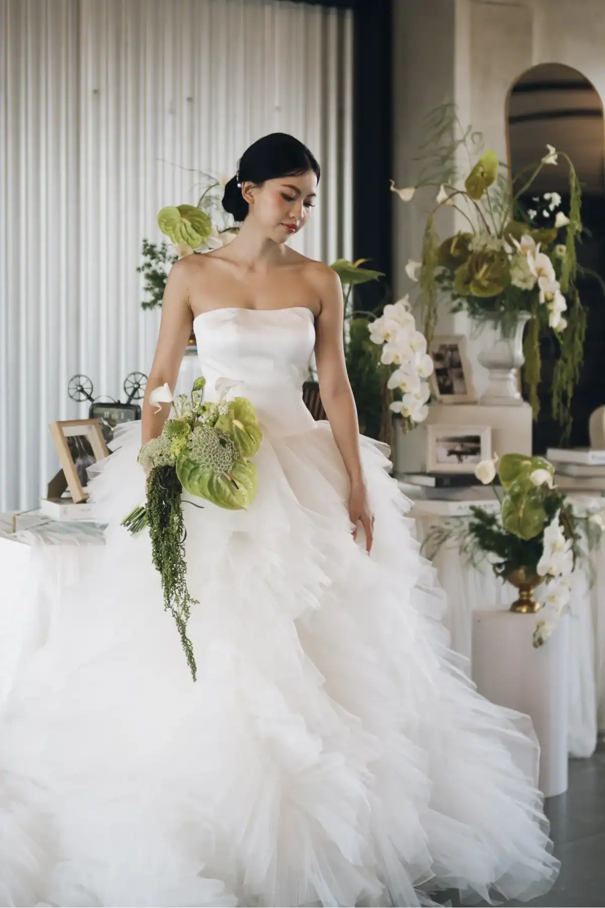 A bride in a strapless white gown holding a bouquet of green and white flowers stands indoors, surrounded by floral arrangements and framed photos.