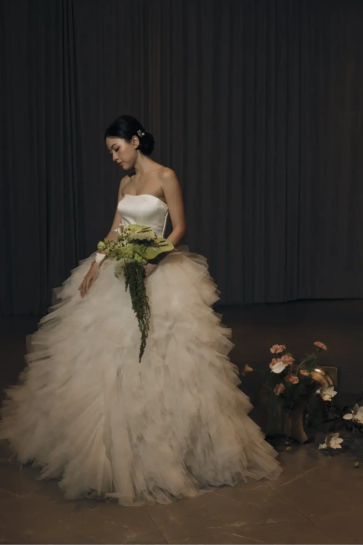 A woman in a strapless, ruffled white wedding dress holds a green bouquet, standing indoors with dim lighting. Flower arrangements sit on the floor nearby.