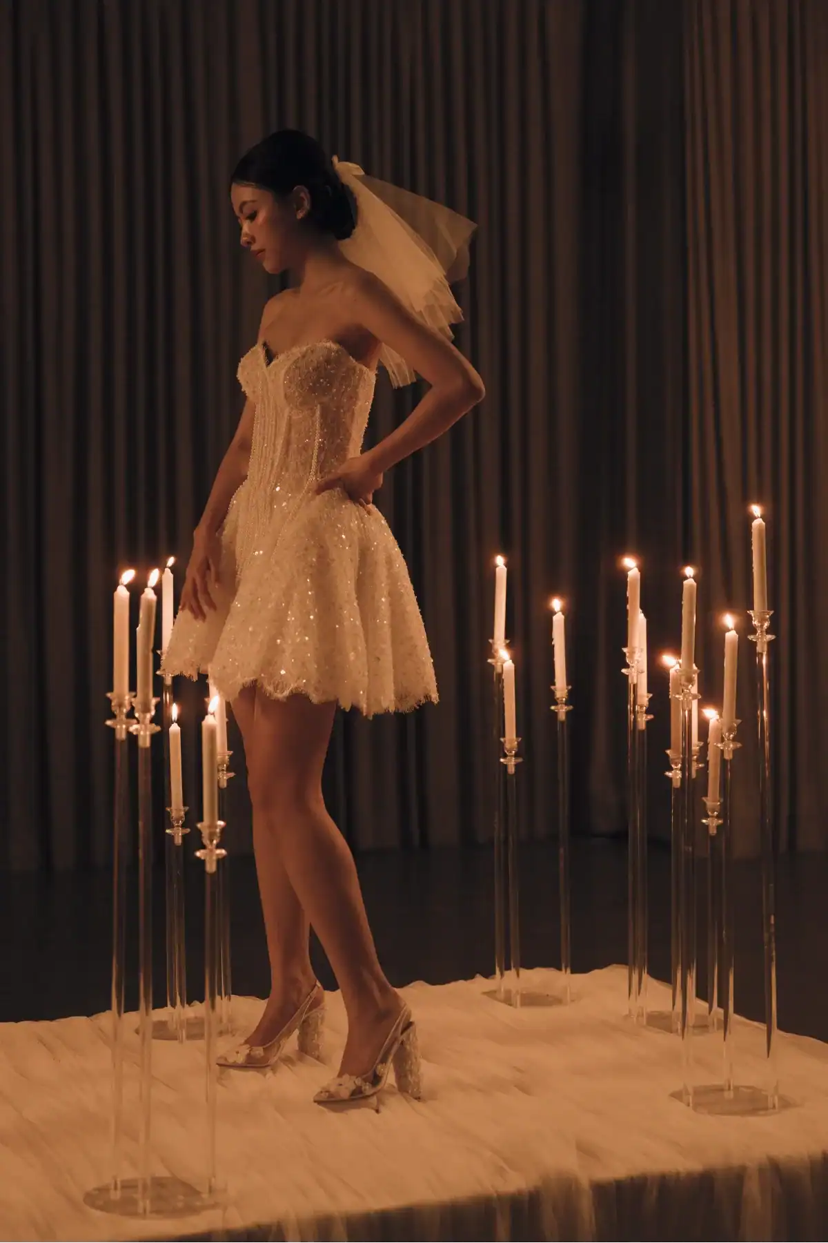 A woman in a short, sparkling white dress and veil stands on a platform surrounded by lit candles on tall stands, with dark curtains in the background.