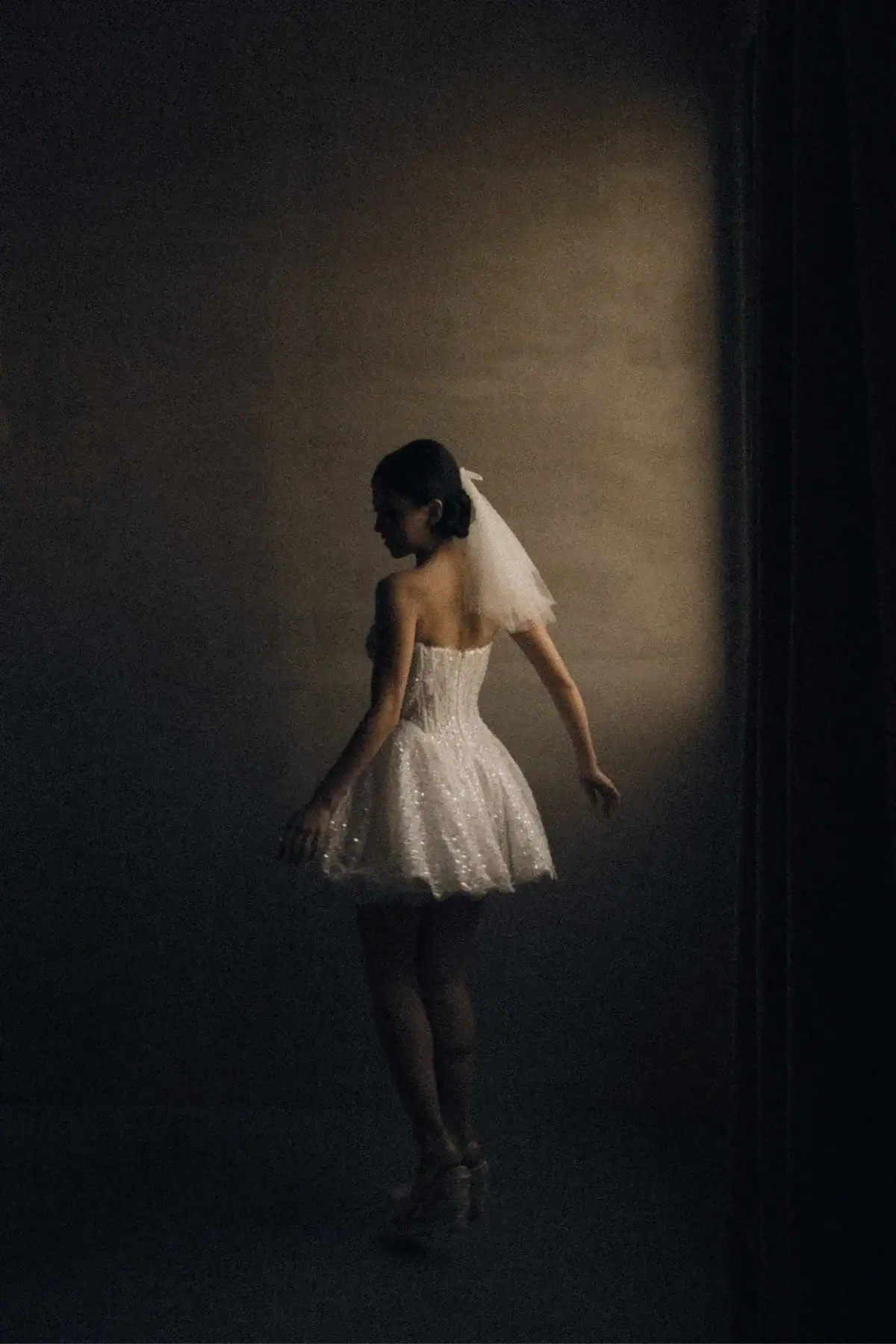 A woman in a short, strapless white dress and veil stands gracefully in a dimly lit room, partially illuminated as she faces away from the camera.