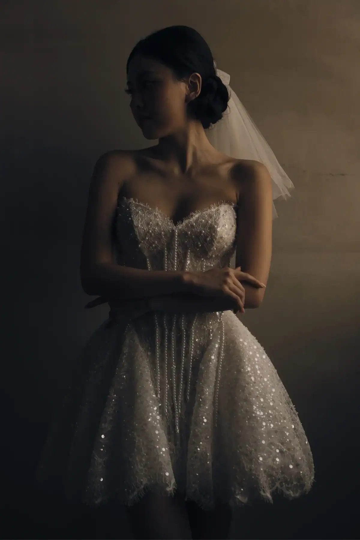 A woman in a sparkly, strapless short white dress with a veil stands against a textured wall, arms folded, looking to the side in soft, moody lighting.